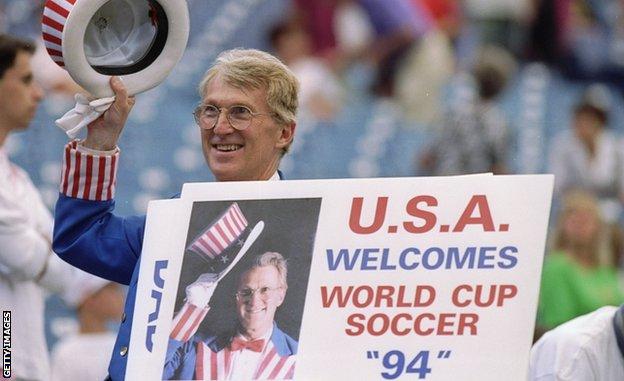 A man with a US-flag themed costume holds a sign announcing the USA welcoming 'World Cup Soccer 94' that also pictures him in the same costume tipping his hat