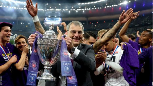 Damien Comolli poses with the French Cup as Toulouse players celebrate behind him