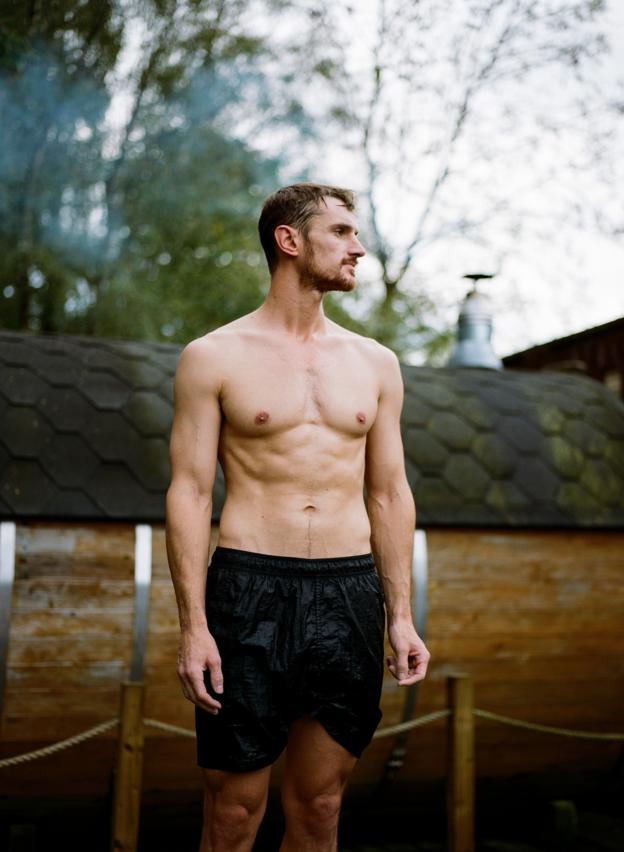 A shirtless man wearing black shorts stands outside a sauna