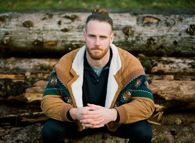 Anthony Mullally, wearing a shearling jacket and clasping his hands, poses in front of logs