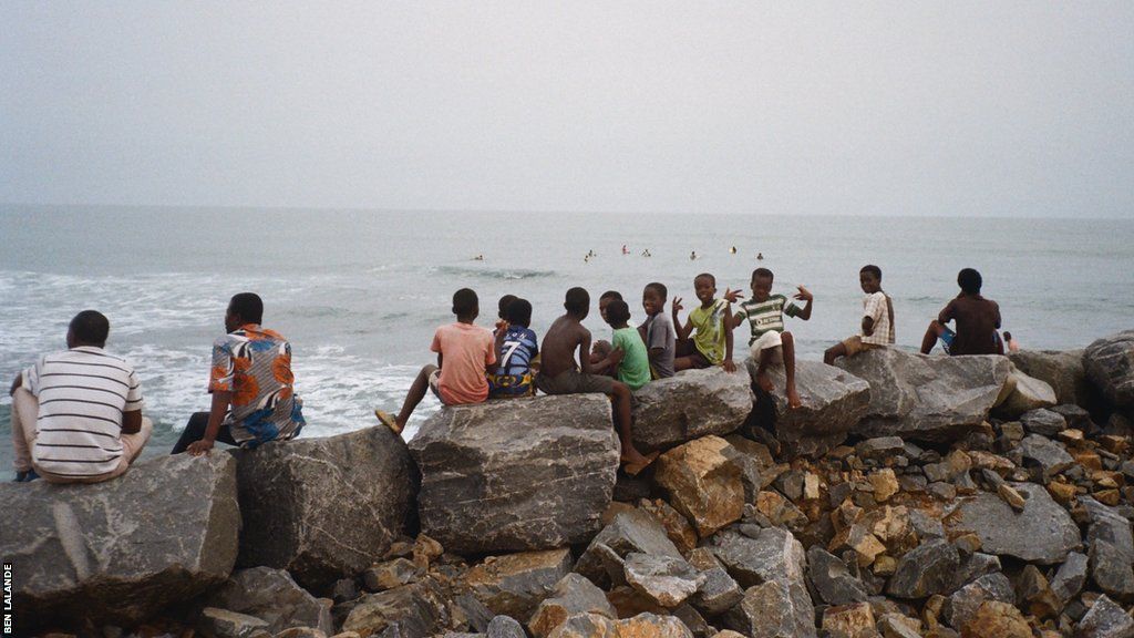 A group of boys and men sit on a breakwater and watch the surfers out at sea