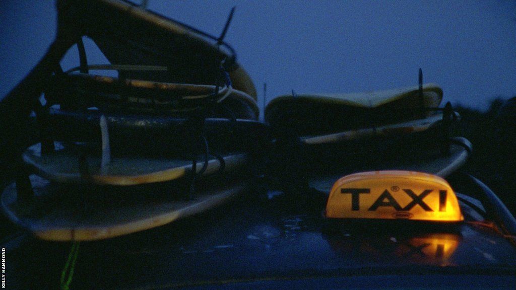 A taxi with illuminated sign is loaded with surfboards on the roof