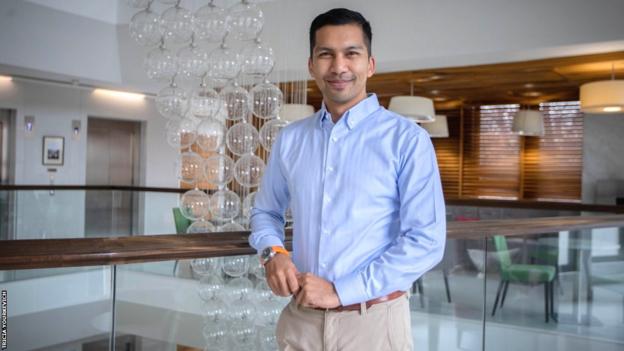 Aron D'Souza smiles while posing near a glass sculpture in an open-plan office