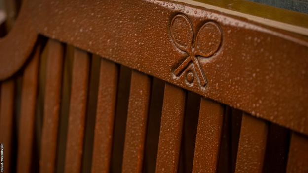 A rain-spattered bench with the Wimbledon logo of two crossed tennis racquets